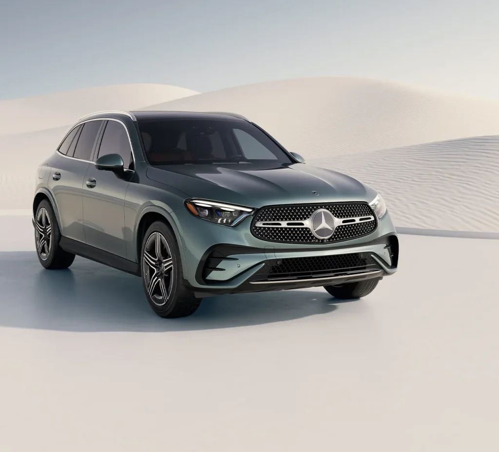 Front angle of a green 2026 Mercedes-Benz GLC parked on a smooth, light-colored surface with sand dunes in the background.