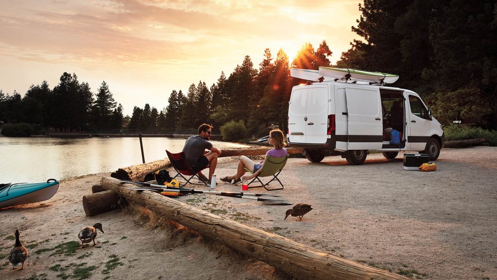 White Mercedes-Benz Sprinter Van parked beside a lakeside campsite with two people sitting in chairs at sunset.
