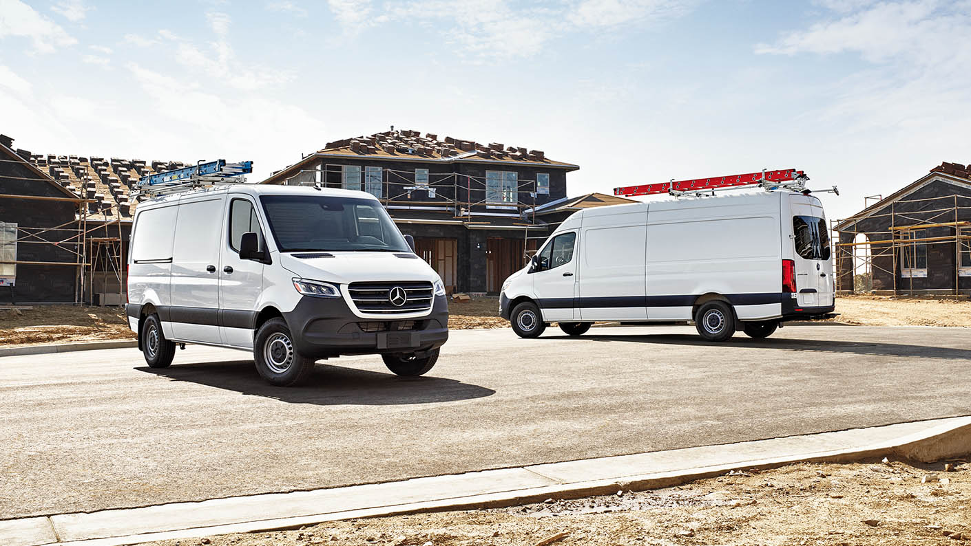 Two white 2026 Mercedes-Benz Sprinter vans parked in front of houses under construction, equipped with ladder racks and work accessories.