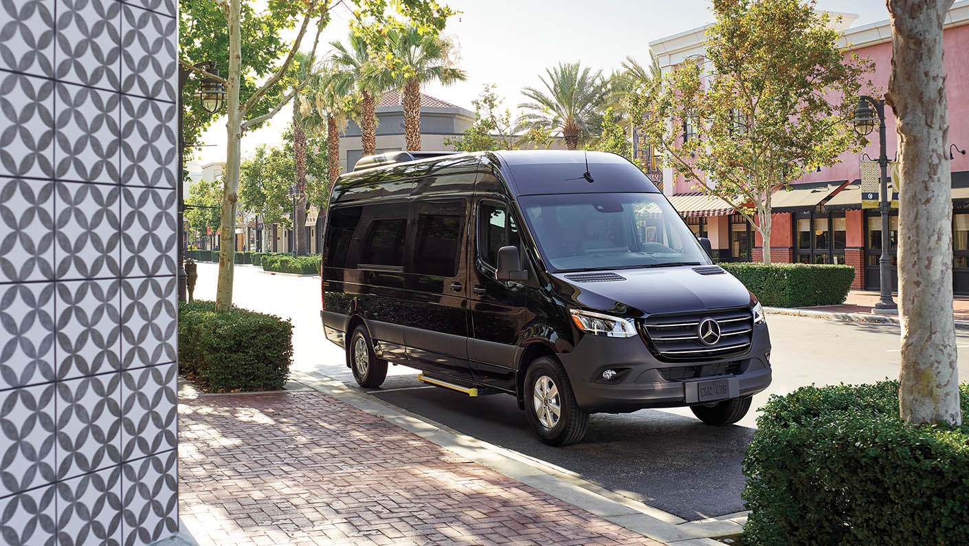 Black Mercedes-Benz Sprinter Van parked on a quiet city street with shops and trees in the background.