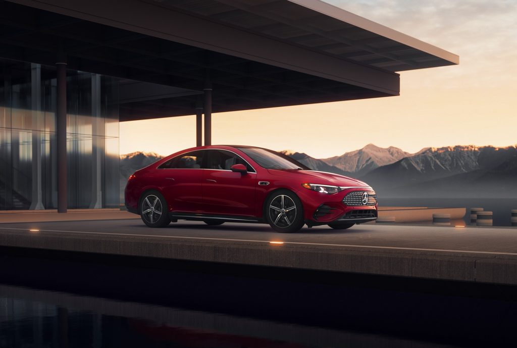 Red all-electric 2026 Mercedes-Benz CLA parked near a modern building with mountains in the background, highlighting luxury sedan styling.