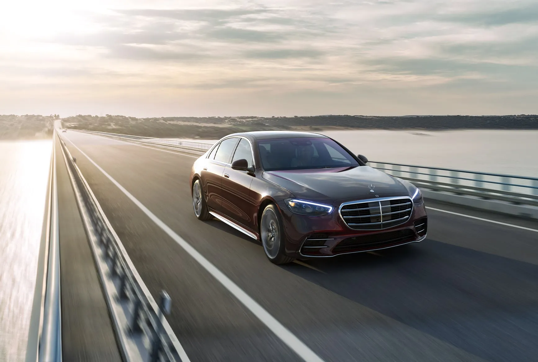 2027 Mercedes-Benz S-Class dark luxury sedan driving on a bridge at sunset.