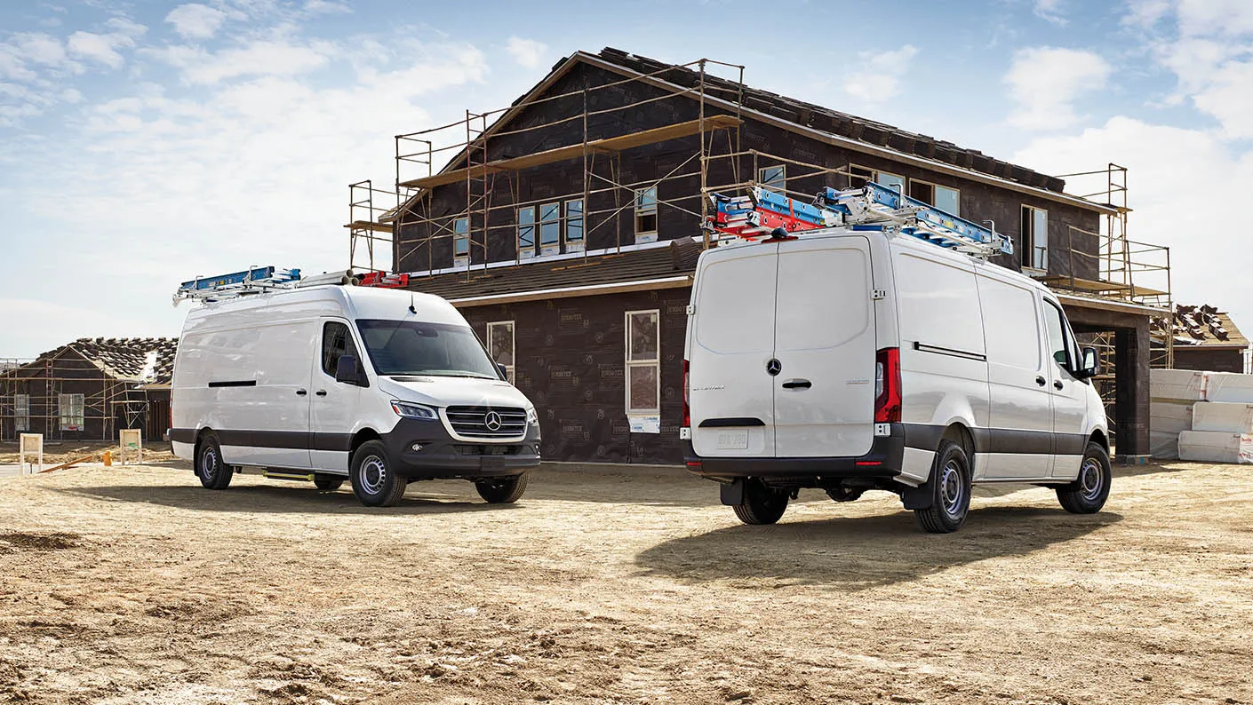 Two Mercedes-Benz Sprinter vans at a construction site equipped with roof-mounted ladders.