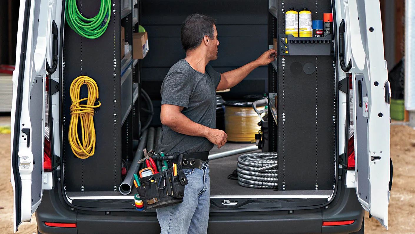 Worker arranging tools and equipment inside a Mercedes-Benz Sprinter cargo van with shelving.