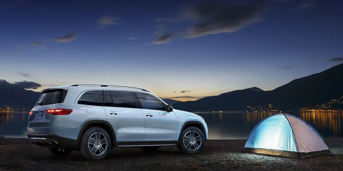 White Mercedes-Benz GLS parked beside a glowing tent near a lake at night with mountains in the distance